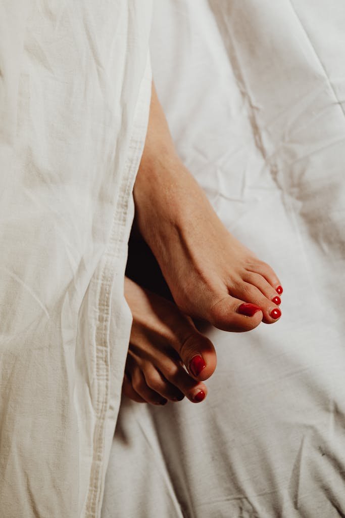 Close-up of feet with red pedicure peeking out from under white sheets, evoking a relaxed and cozy morning vibe.